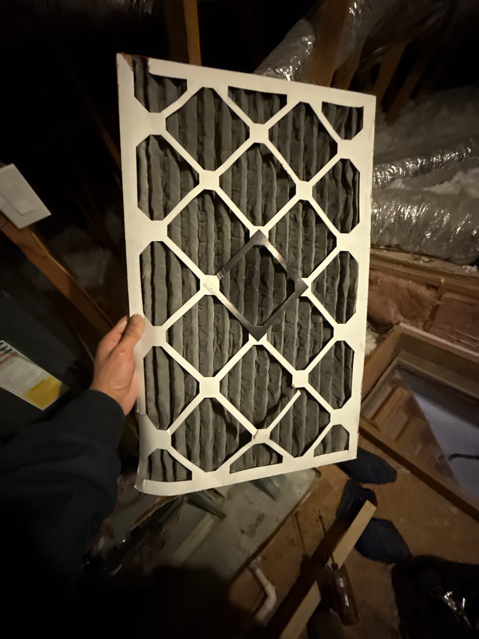 Hand holding up a used pleated air filter caked with dark gray dust, in an attic next to an air handler