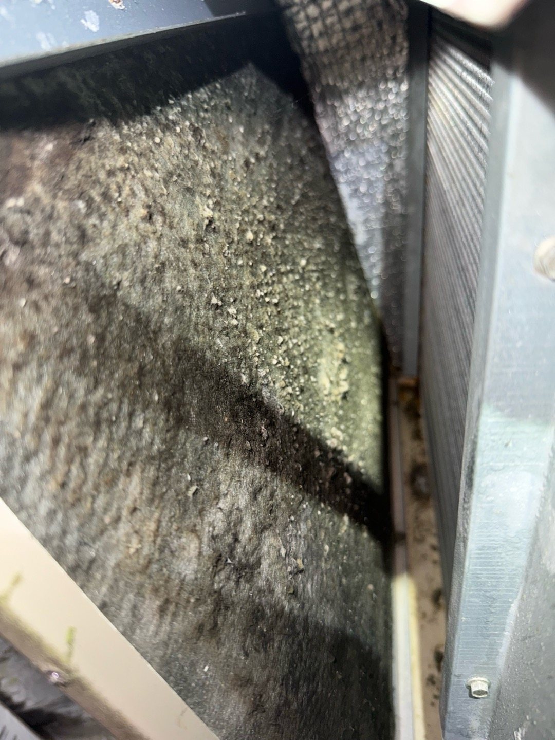 A clogged furnace air filter still in its filter slot, matted gray with dust and pulling inward from the suction of the blower