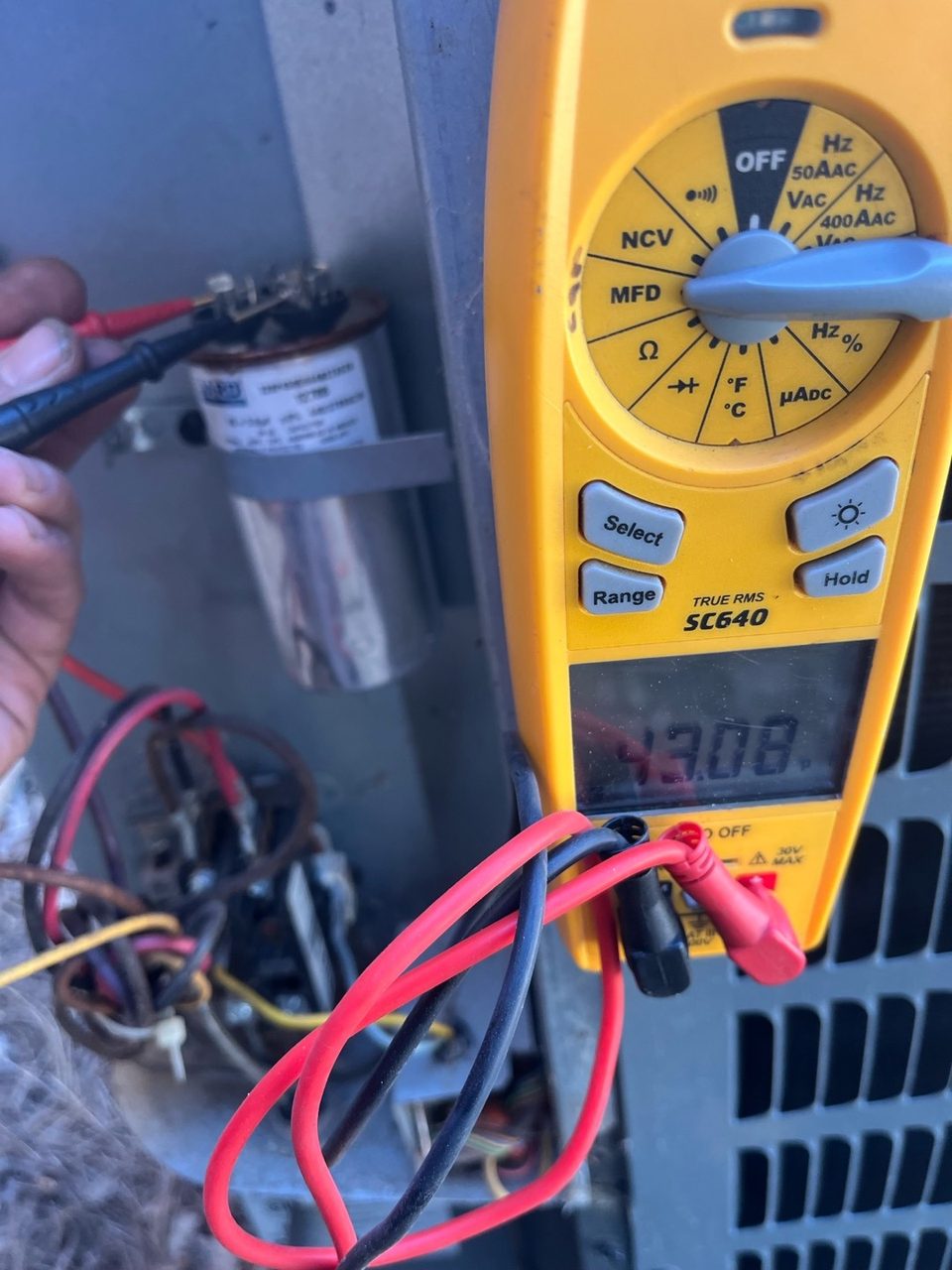 Technician using a yellow digital multimeter to test the capacitor inside an outdoor AC unit; the meter reads 43.08 microfarads