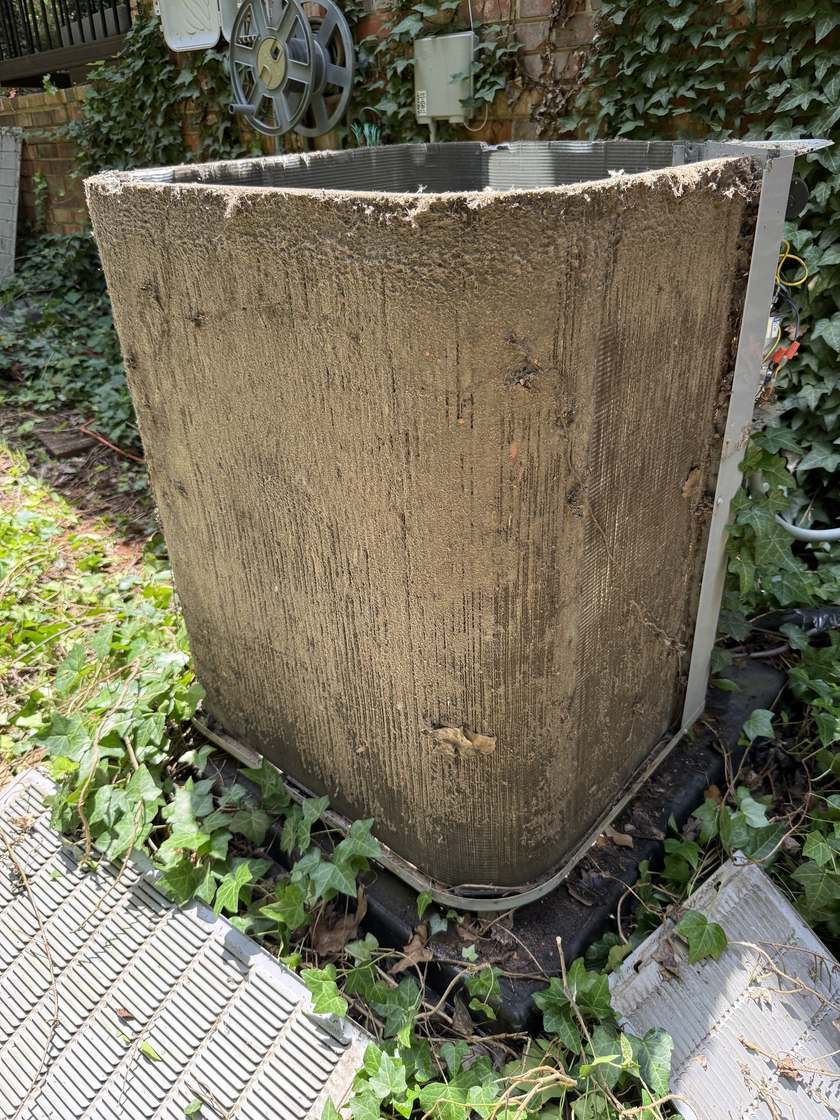 Outdoor air conditioner condenser with its panels removed, the coil fins covered in a thick matted layer of dust and debris, beside a brick wall with ivy at its base