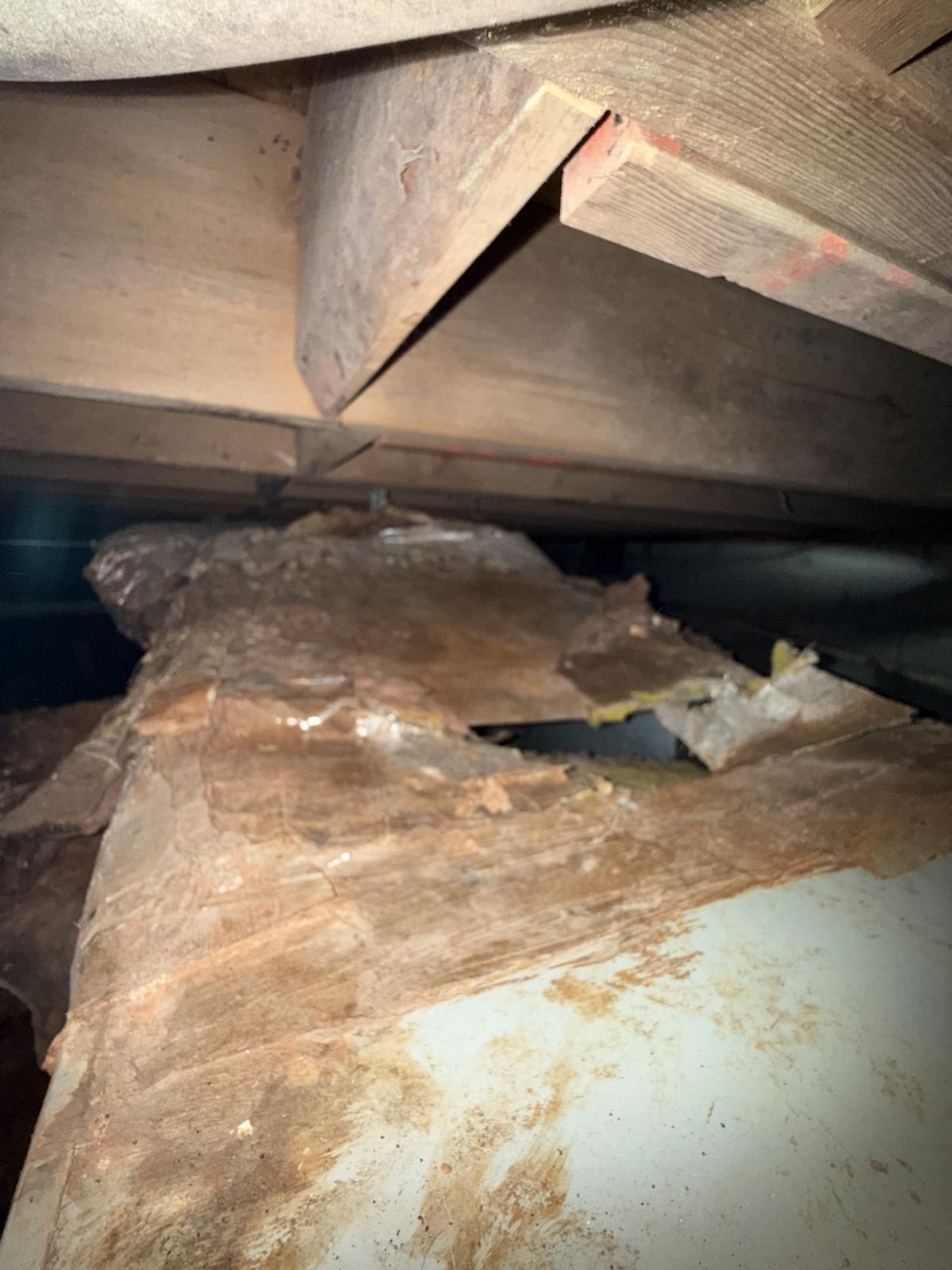Torn, stained duct wrap with a large ragged hole in a dark crawl space beneath wooden floor joists