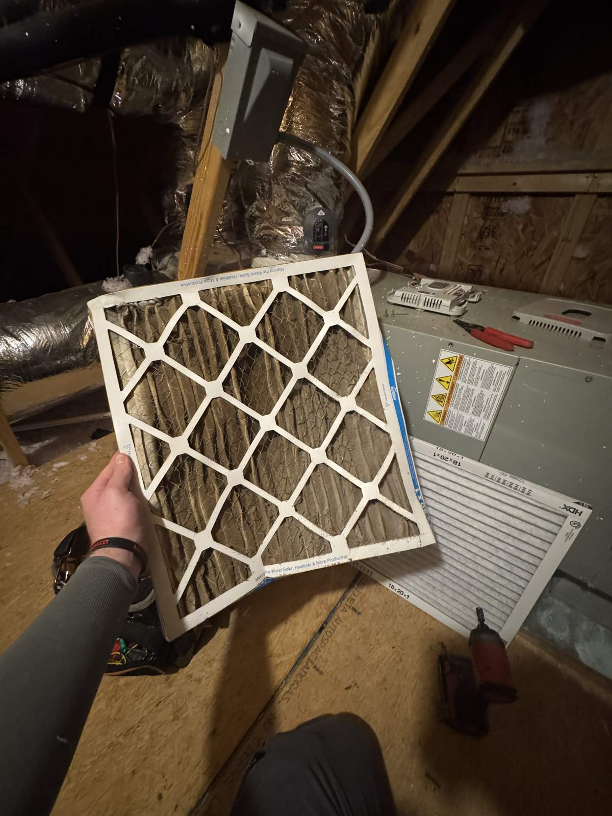 Hand holding a dirty, dust-clogged pleated air filter with a bent frame beside a clean new filter on an attic floor