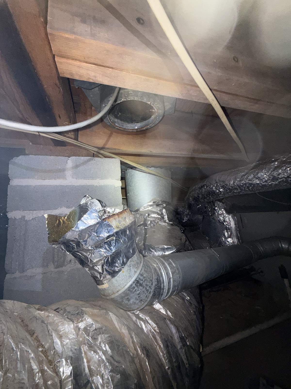 Metal pipe elbow wrapped in crumpled silver foil tape below an open round vent collar between floor joists in a crawl space