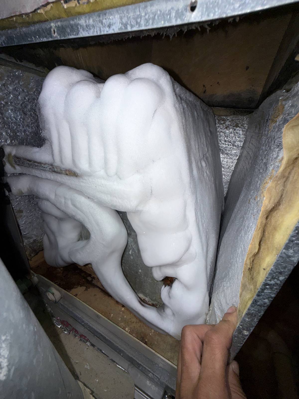 Evaporator coil and copper lines inside an air handler covered in thick ice, with a hand pulling back foil-faced insulation