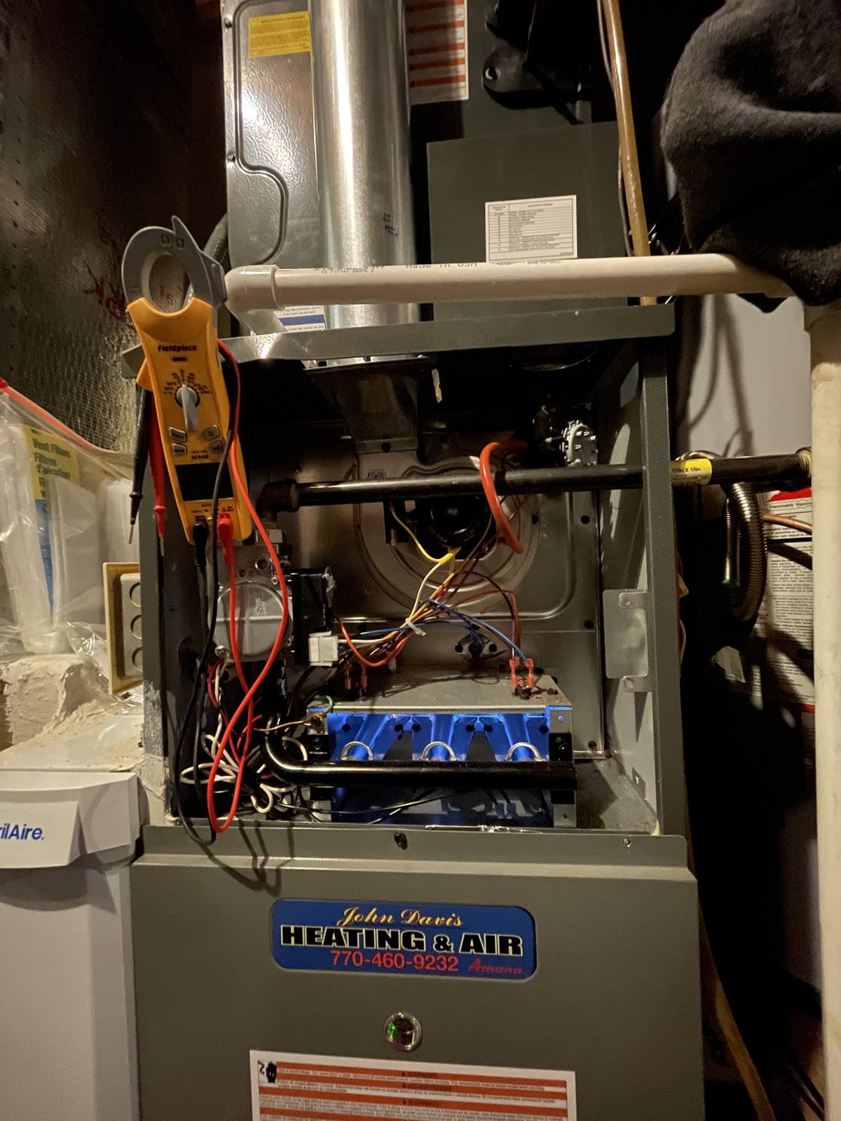 Gas furnace with the upper front panel removed and four blue burner flames lit, with a yellow multimeter hanging from the cabinet and a service-company sticker on the lower panel