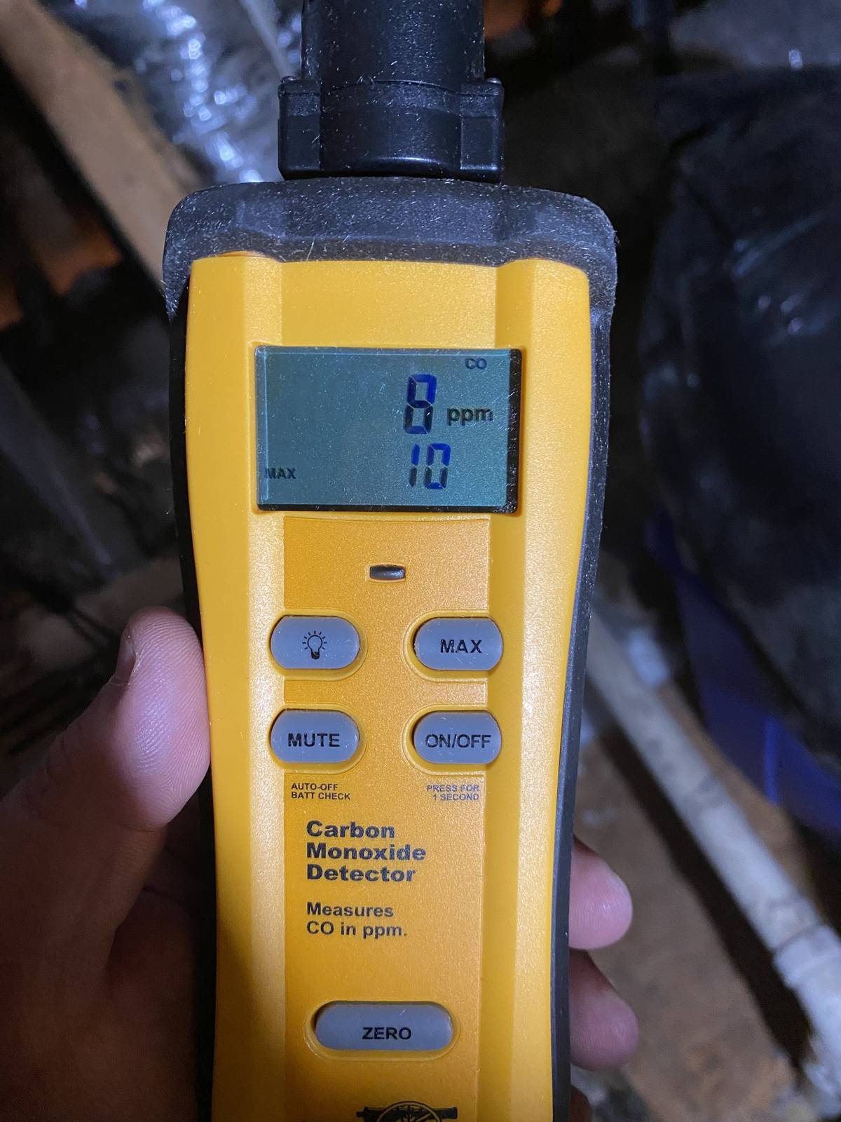 Hand holding a yellow handheld carbon monoxide detector whose display reads 8 ppm, with a maximum of 10, in a dim attic or crawl space