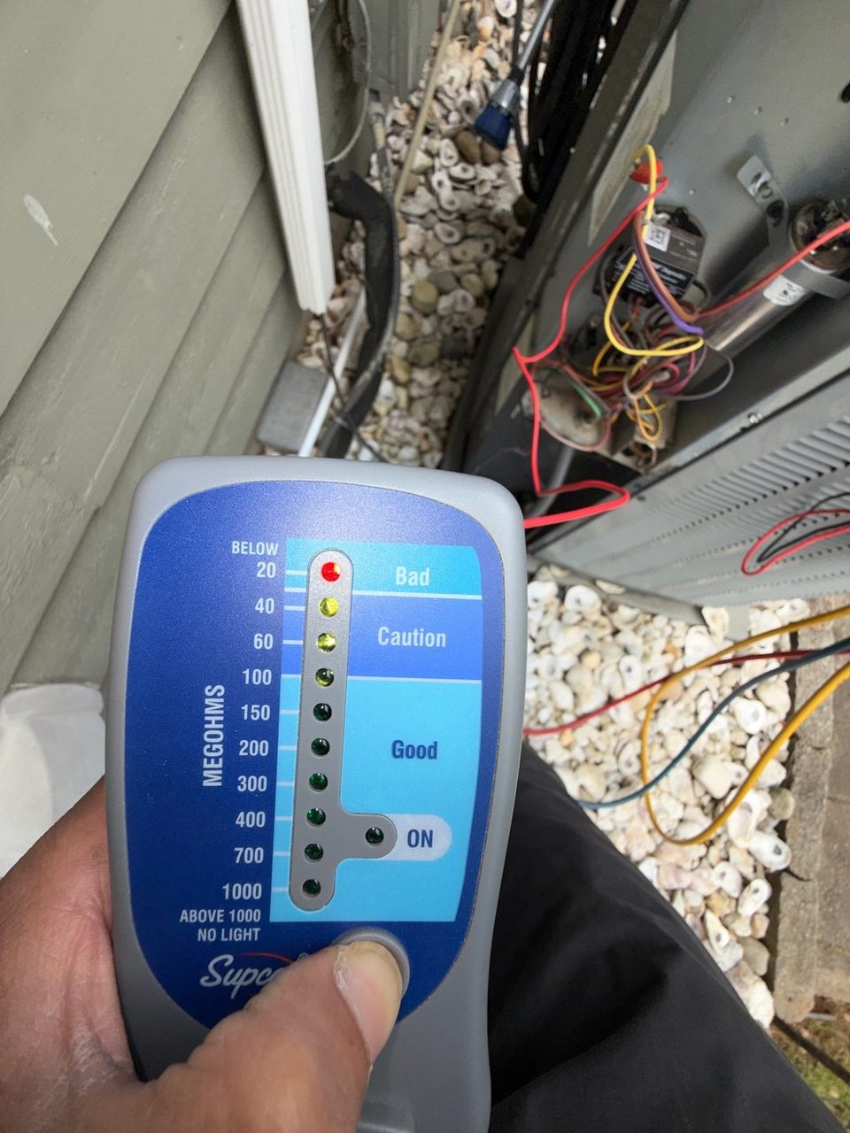 Technician holding a handheld insulation (megohm) tester in front of the open electrical compartment of an outdoor AC unit