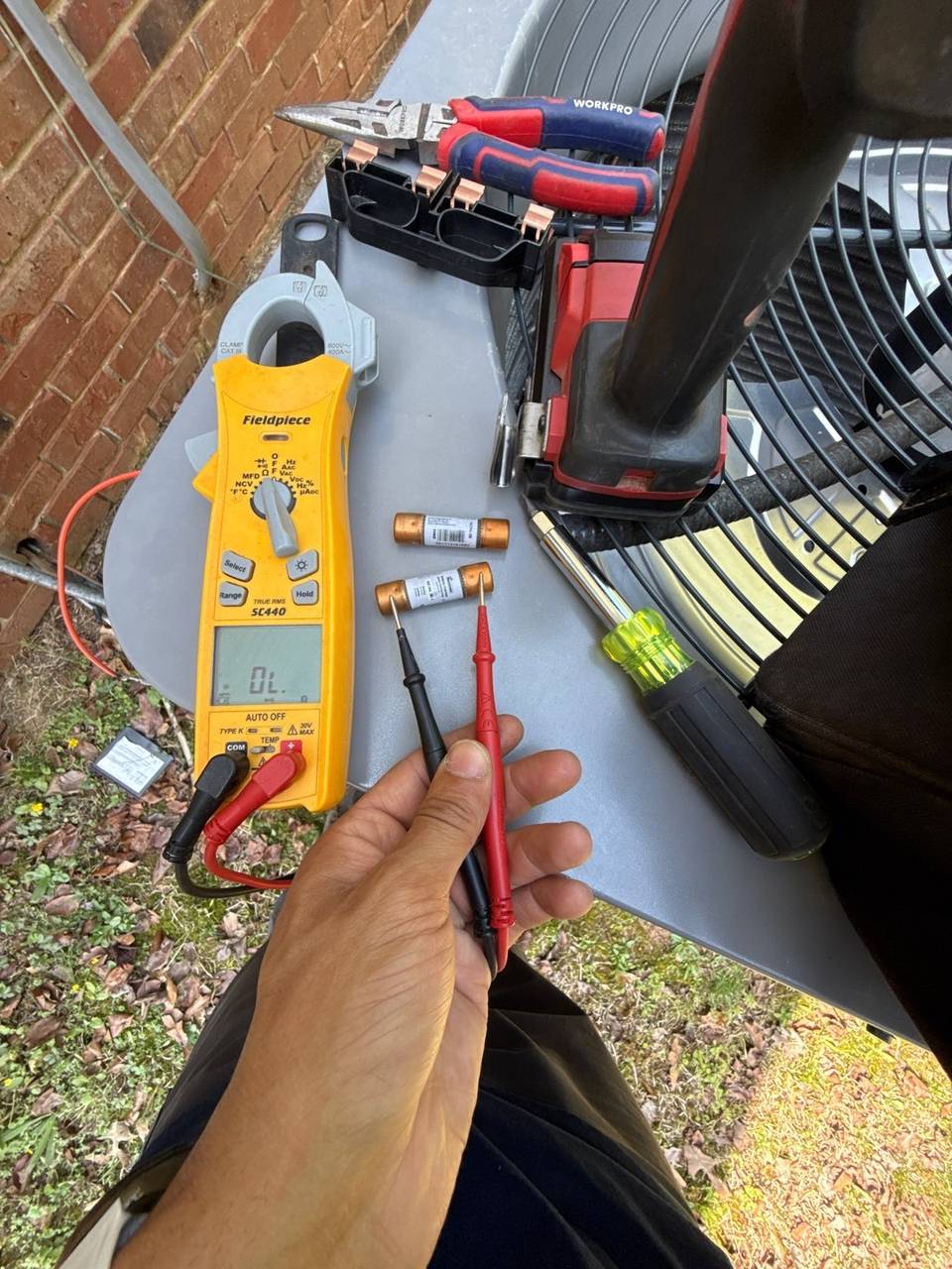Multimeter and probes testing two small cartridge fuses laid on top of an outdoor AC unit, with hand tools beside them