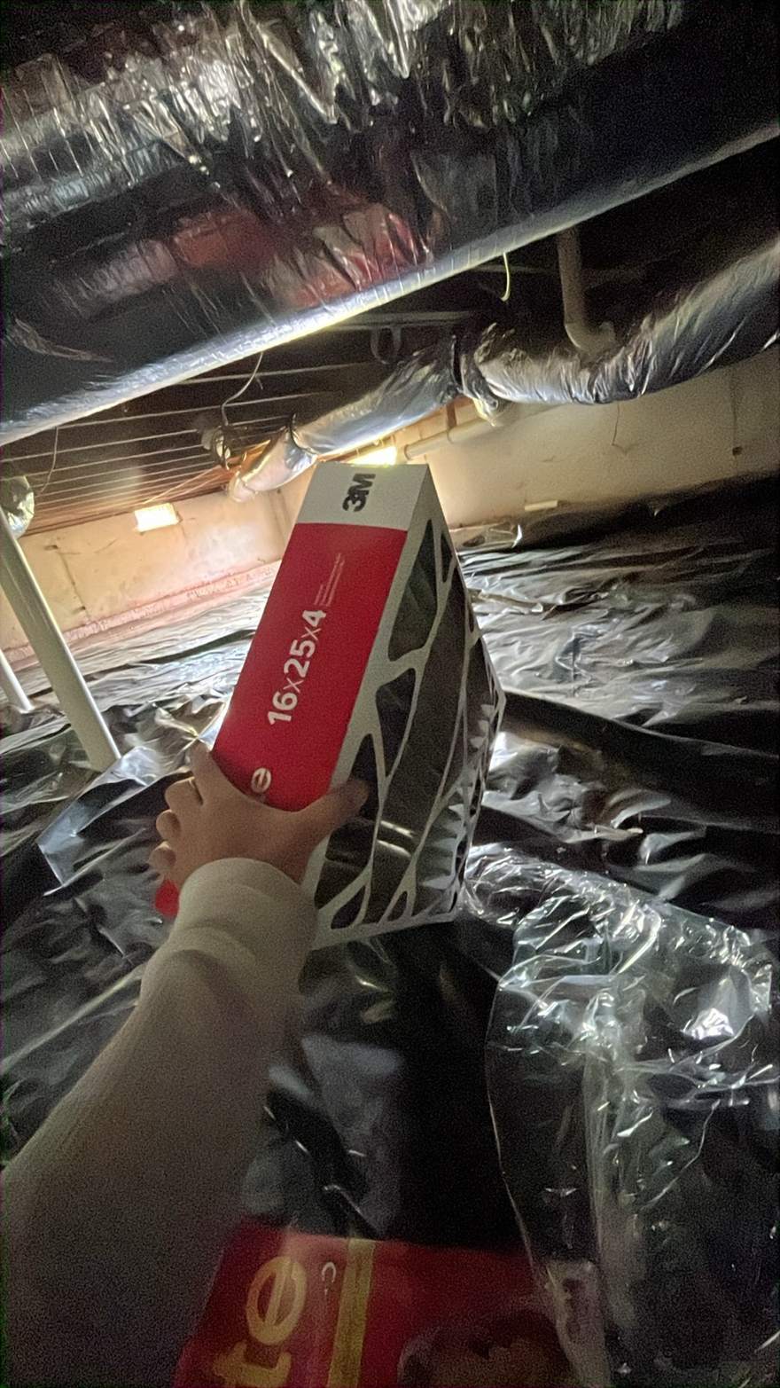 Hand holding a new 16x25x4 pleated air filter in a crawlspace next to insulated ductwork