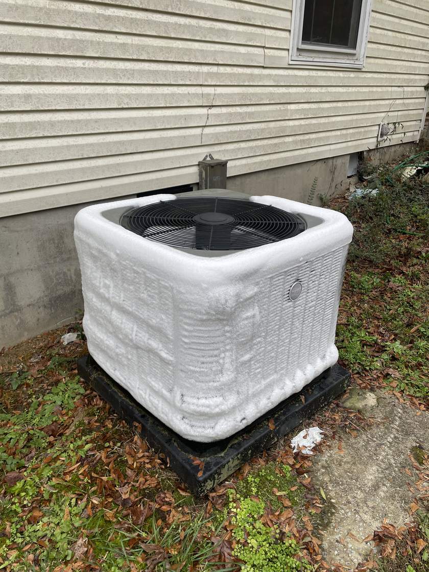 Outdoor air conditioner condenser unit beside a house, its side panels covered in a thick layer of white frost and ice, sitting on a black pad