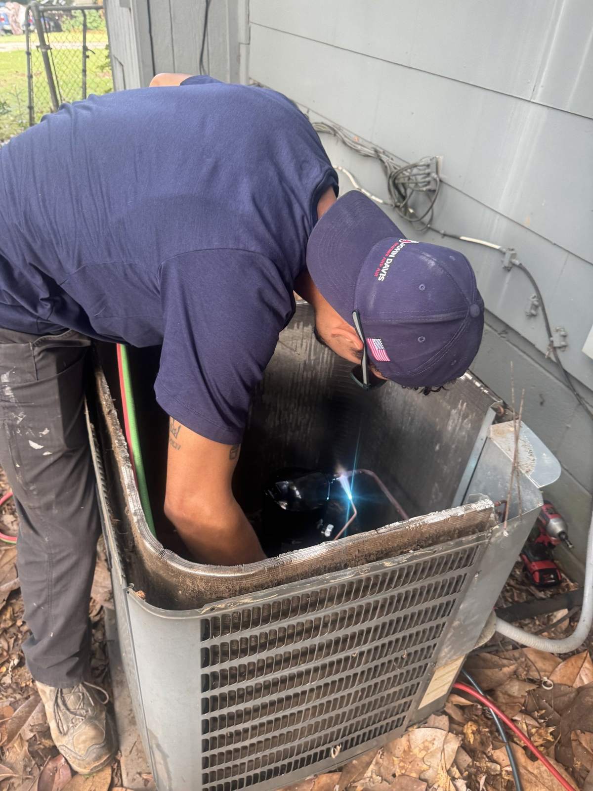 Technician in a navy cap and short-sleeved shirt leaning down into an open outdoor air conditioner beside a gray-sided house, with a cordless drill and dry leaves on the ground