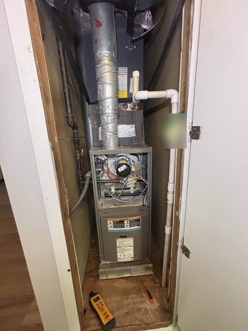 Upflow gas furnace in a hallway closet with the burner panel removed, showing the flue pipe, gas line and condensate drain during a heating inspection