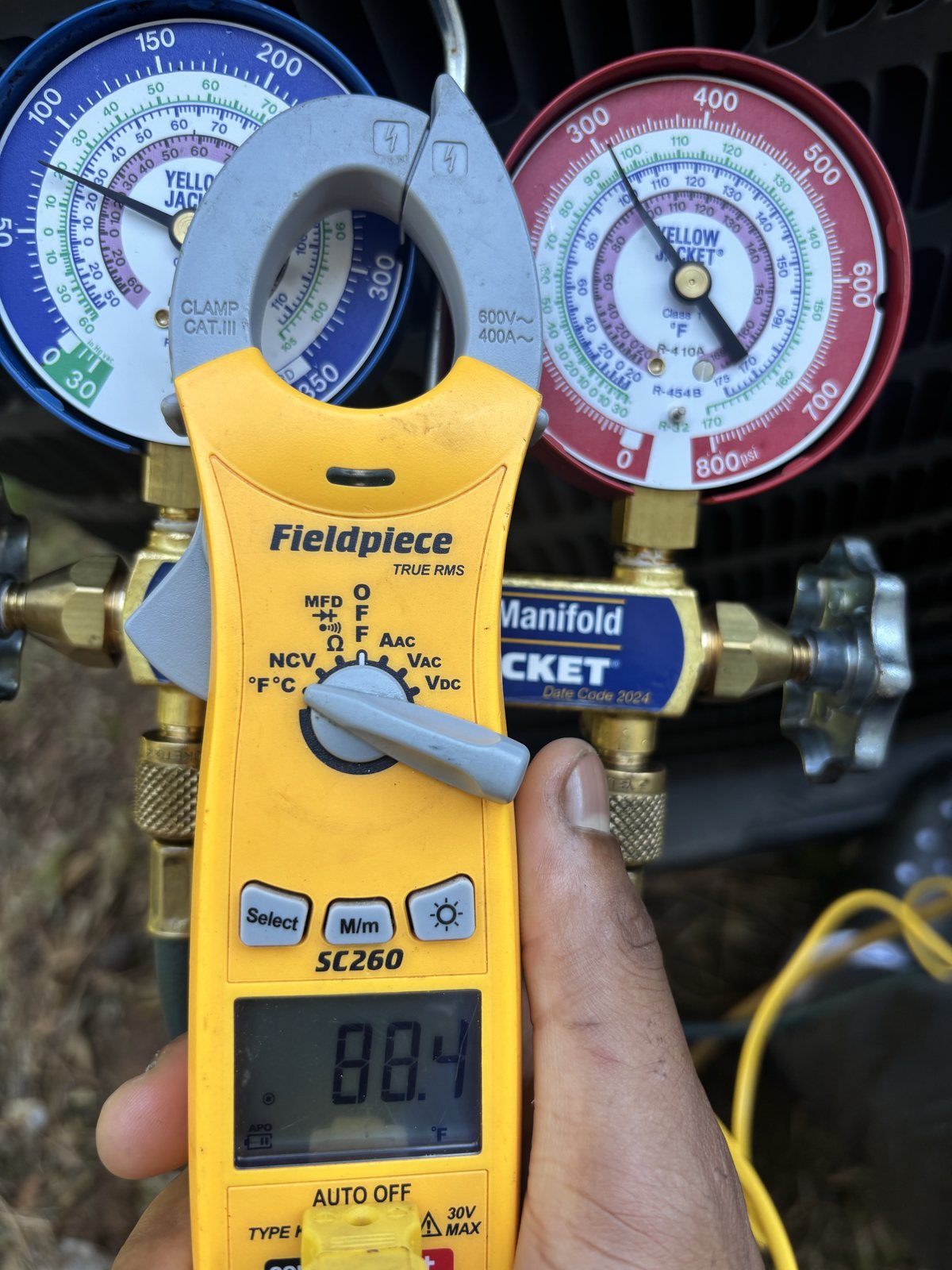 Technician holding a clamp meter in front of refrigerant gauges while diagnosing an air conditioner that won't cool